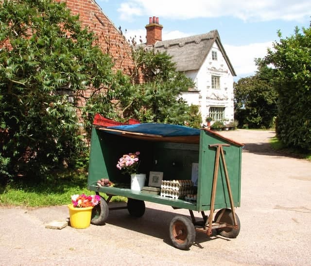 A green farm trailer parked outside a thatched cottage, its open side stacked with trays of eggs and a jug of cut flowers