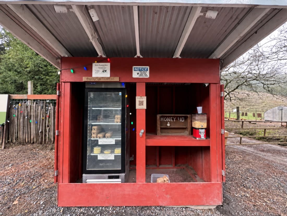 A red timber honesty stand at the end of a driveway, with a glass-fronted cooler of eggs and a box of honey on the counter